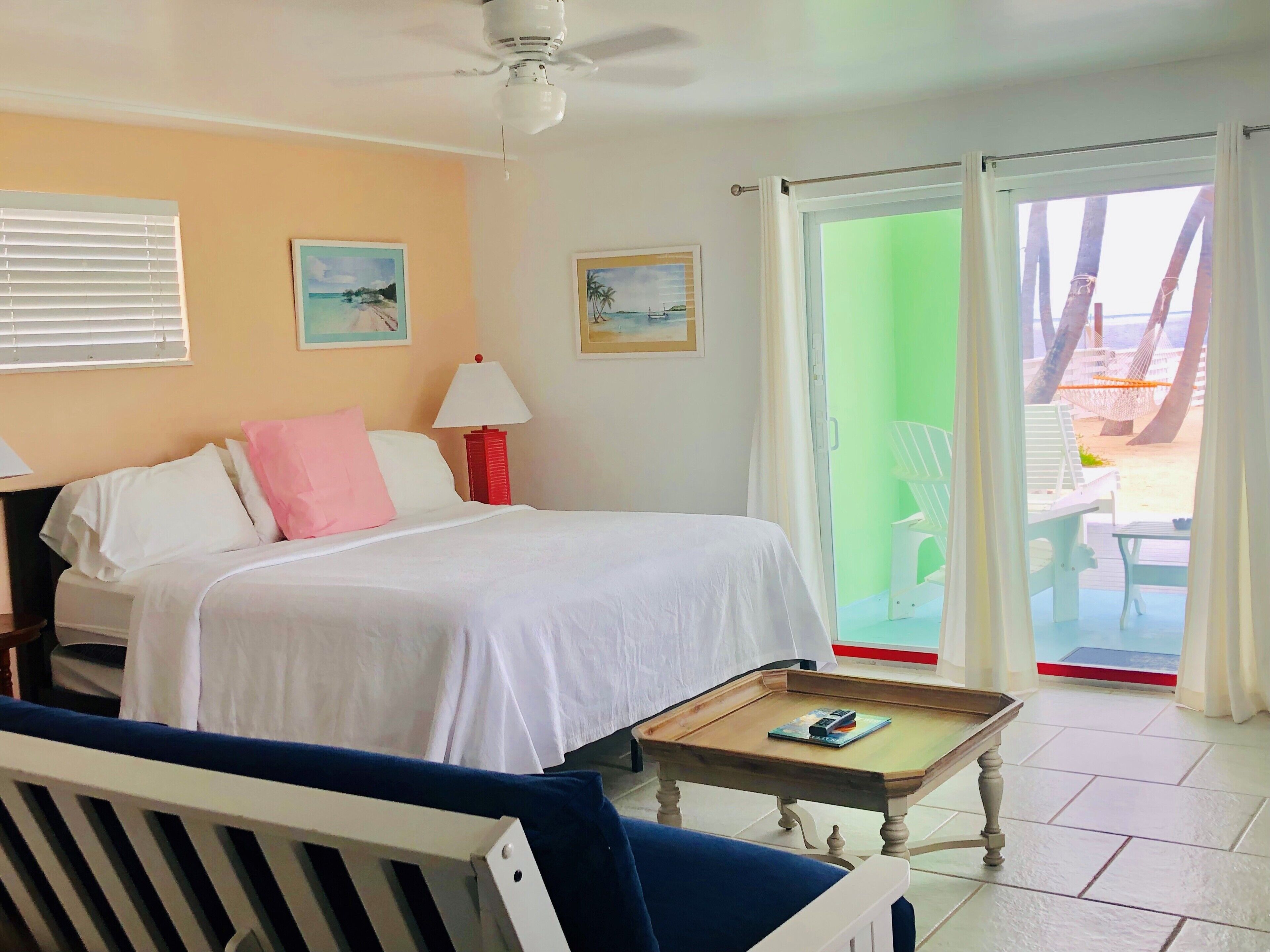 A hotel room with a bed, a nightstand, a lamp, a small table, and a view of the beach through the sliding glass door.