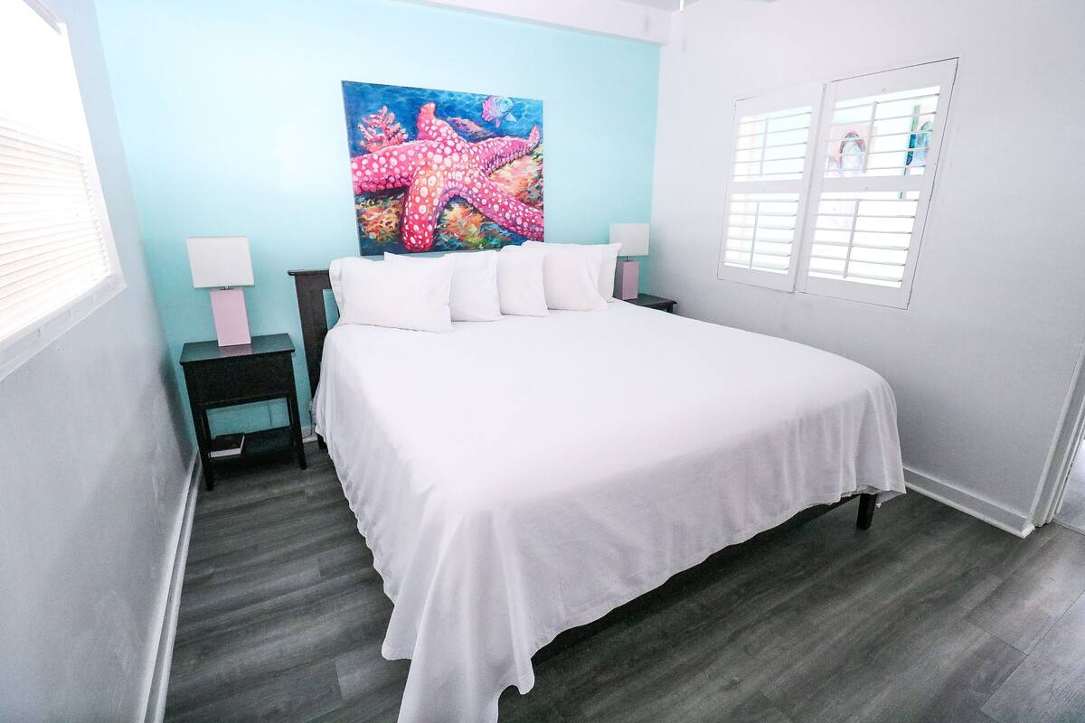 A bedroom with a large bed, a nightstand with a lamp, a painting of a starfish on the wall, and a window with blinds.
