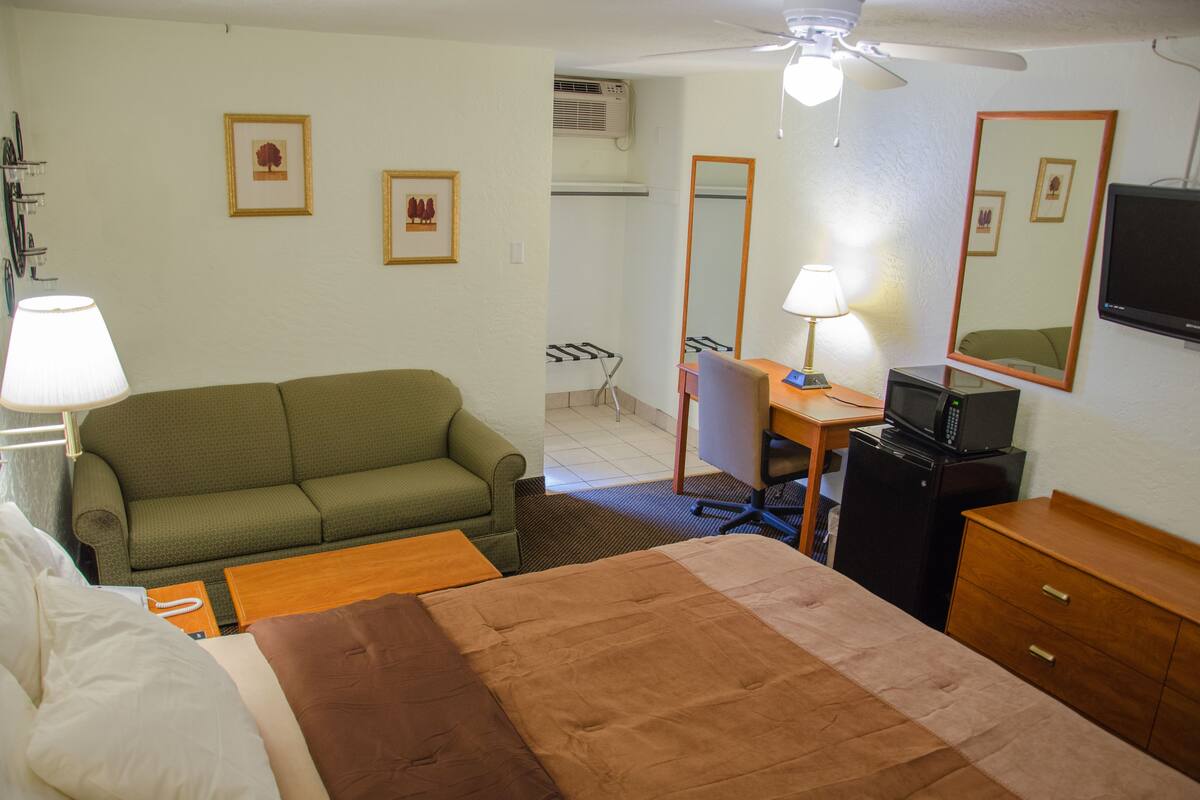 A hotel room with a green sofa, a wooden desk with a TV, a mirror, and a ceiling fan.