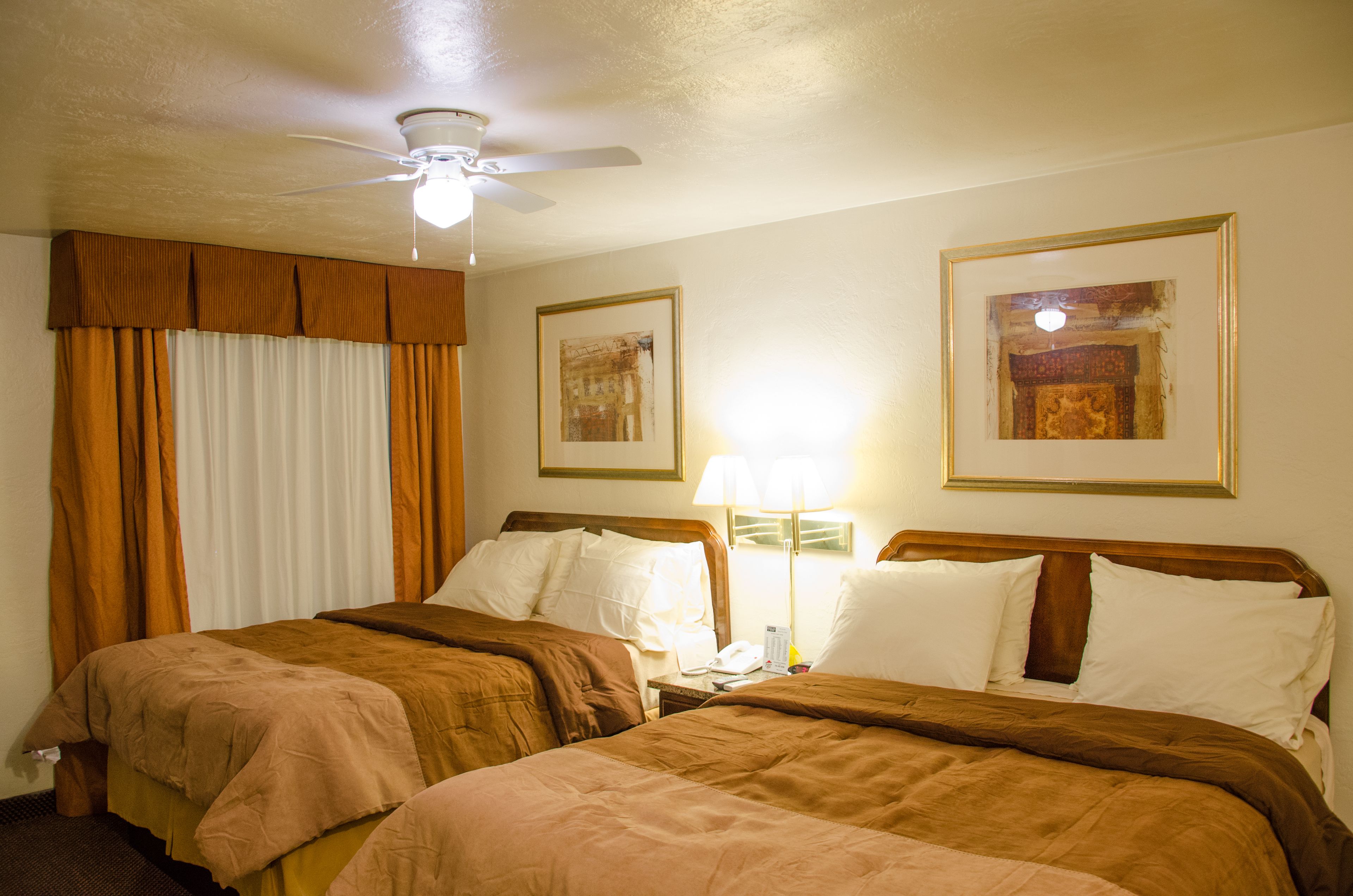 A hotel room with two beds, a ceiling fan, a nightstand with a lamp, and framed artwork on the walls.