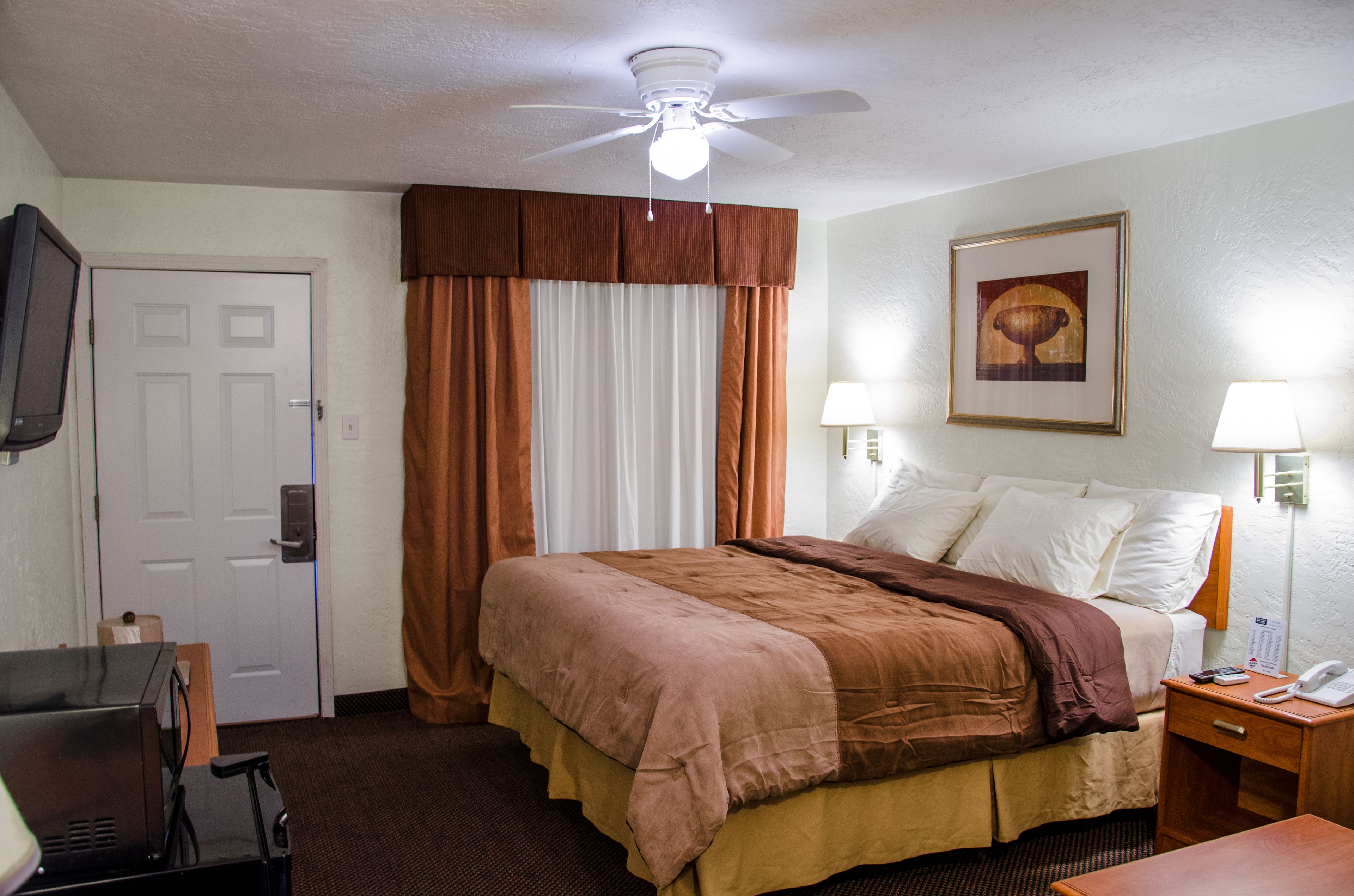 A hotel room with a bed, a desk, a chair, a ceiling fan, a wall-mounted TV, and a painting.