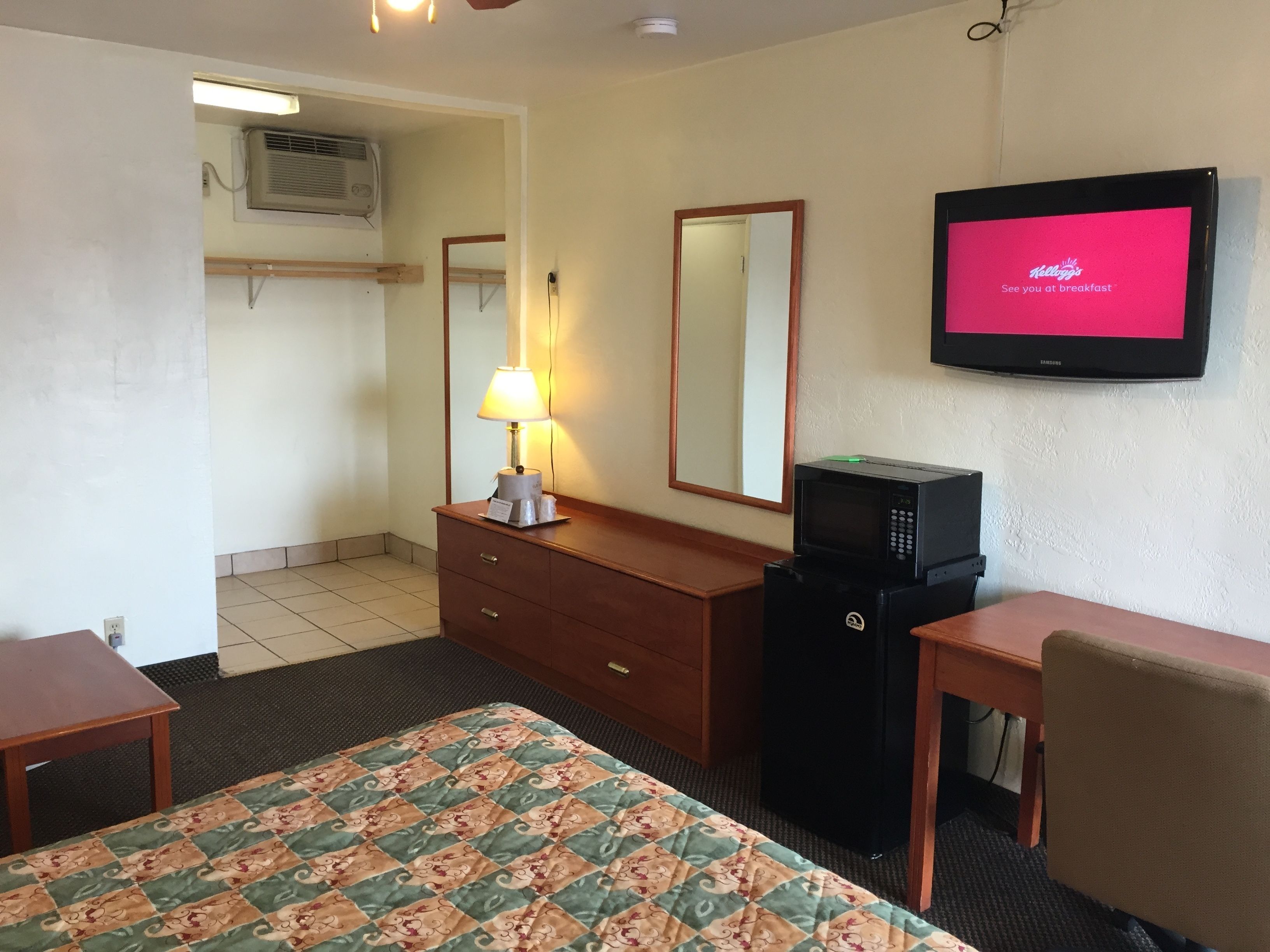 A hotel room with a bed, a desk, a dresser, a microwave, a television displaying a breakfast message, a mirror, and an air conditioning unit.