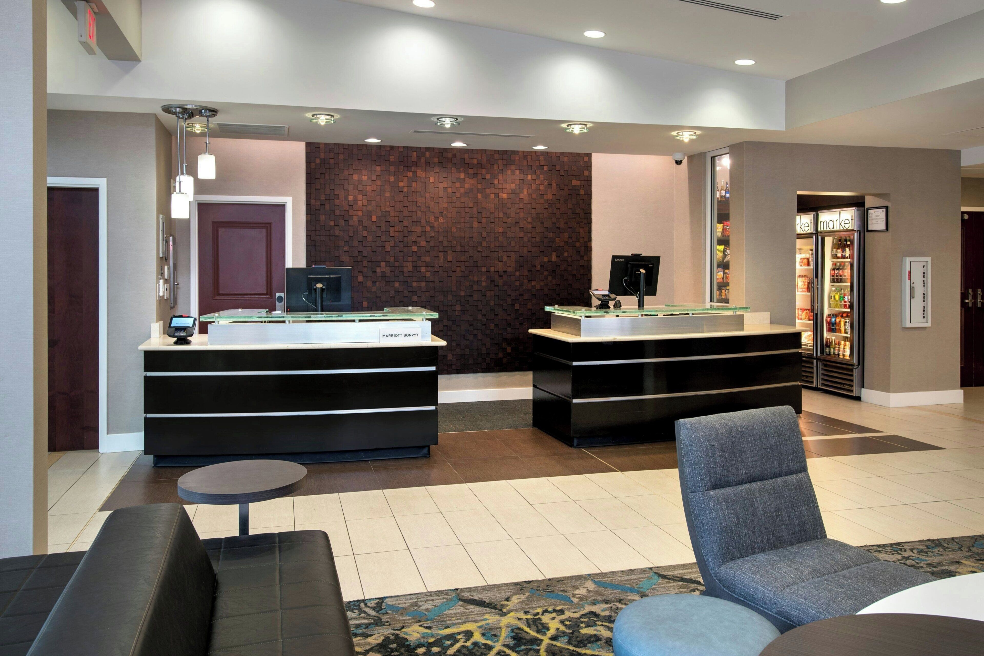 A hotel reception area with two reception desks, a seating area with a sofa and chairs, and a refrigerator stocked with beverages.