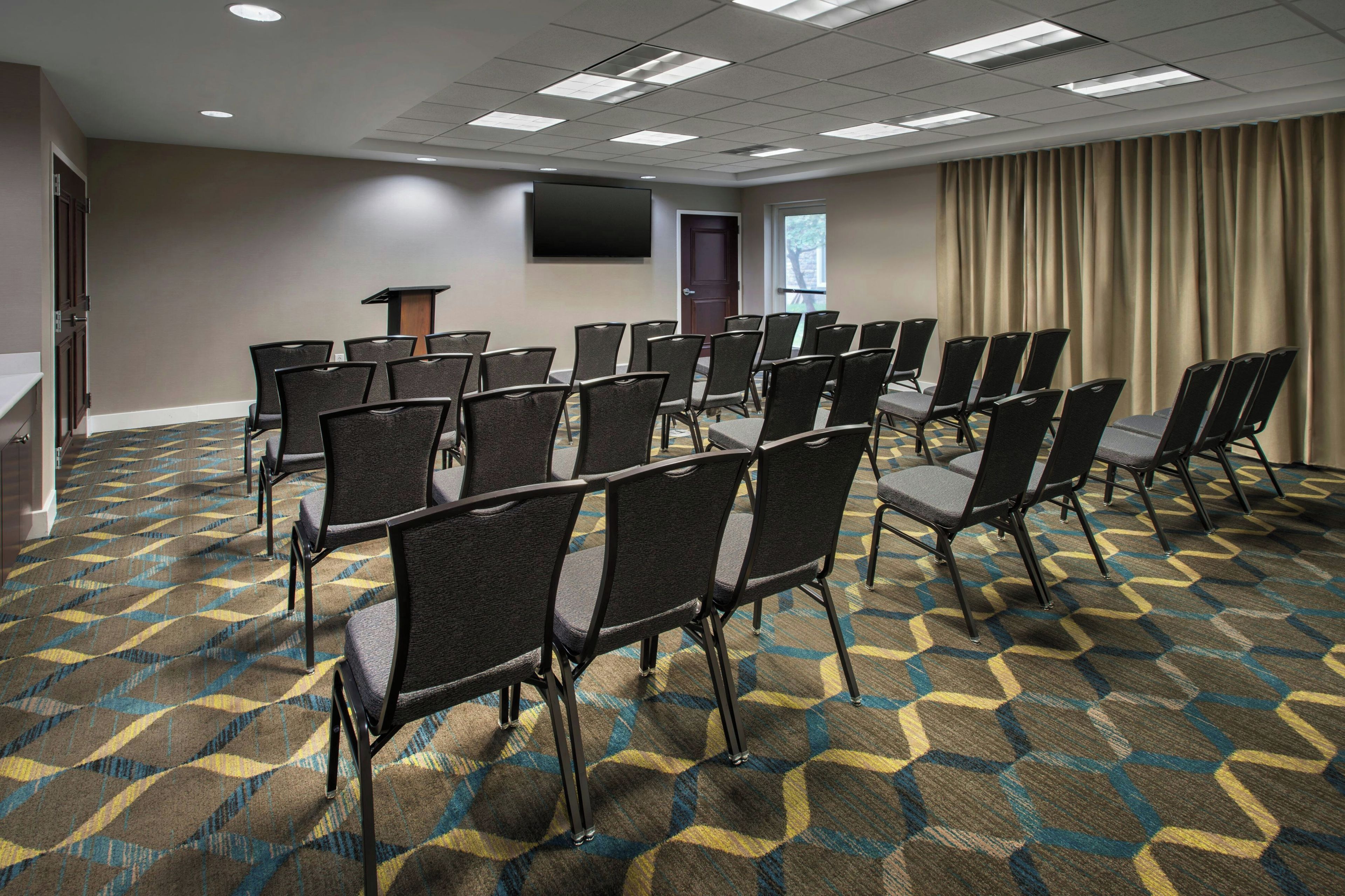 A conference room with rows of chairs, a podium, a flat-screen TV, and a patterned carpet.