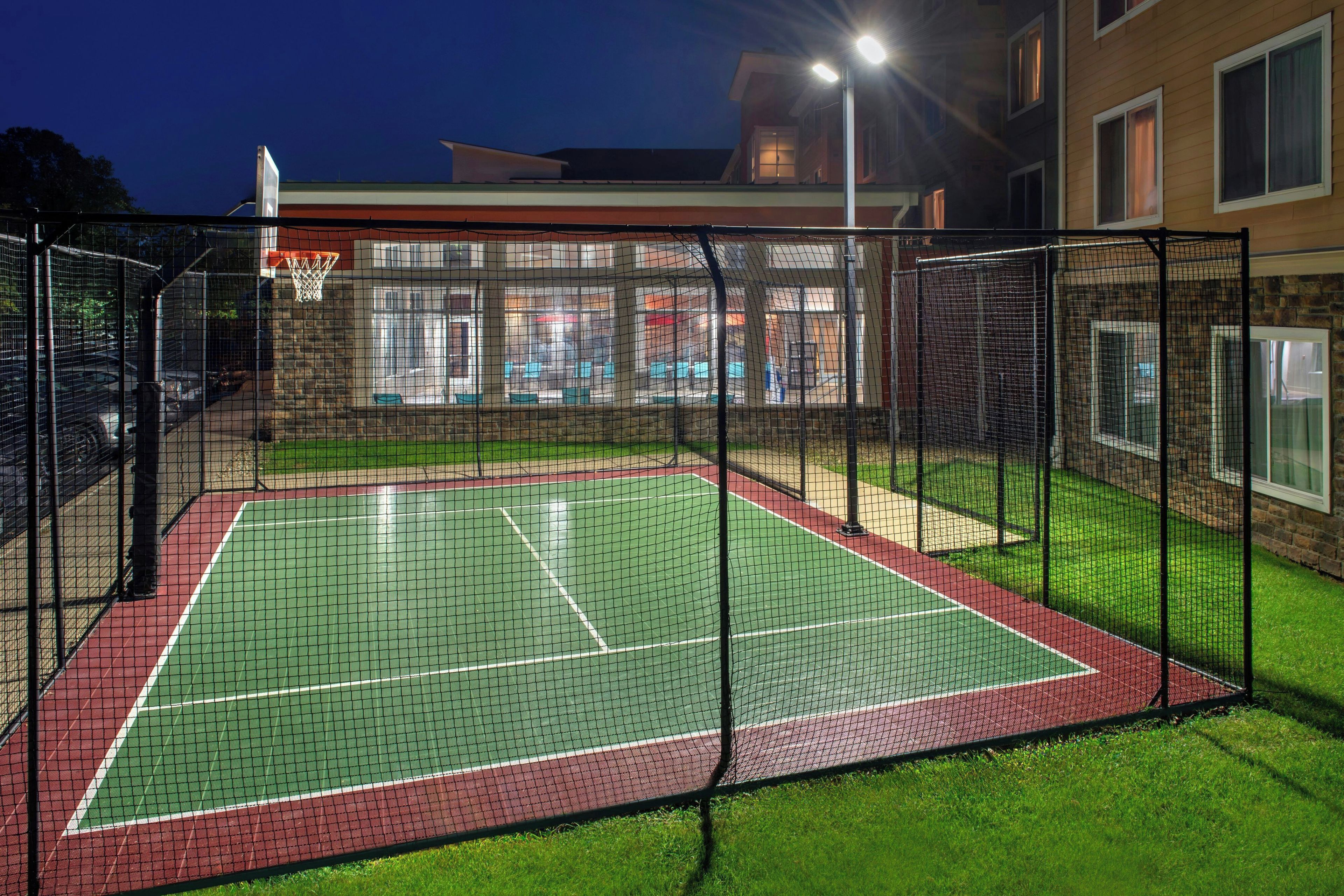 An outdoor tennis court with a net, surrounded by a fence, and a building with windows in the background.