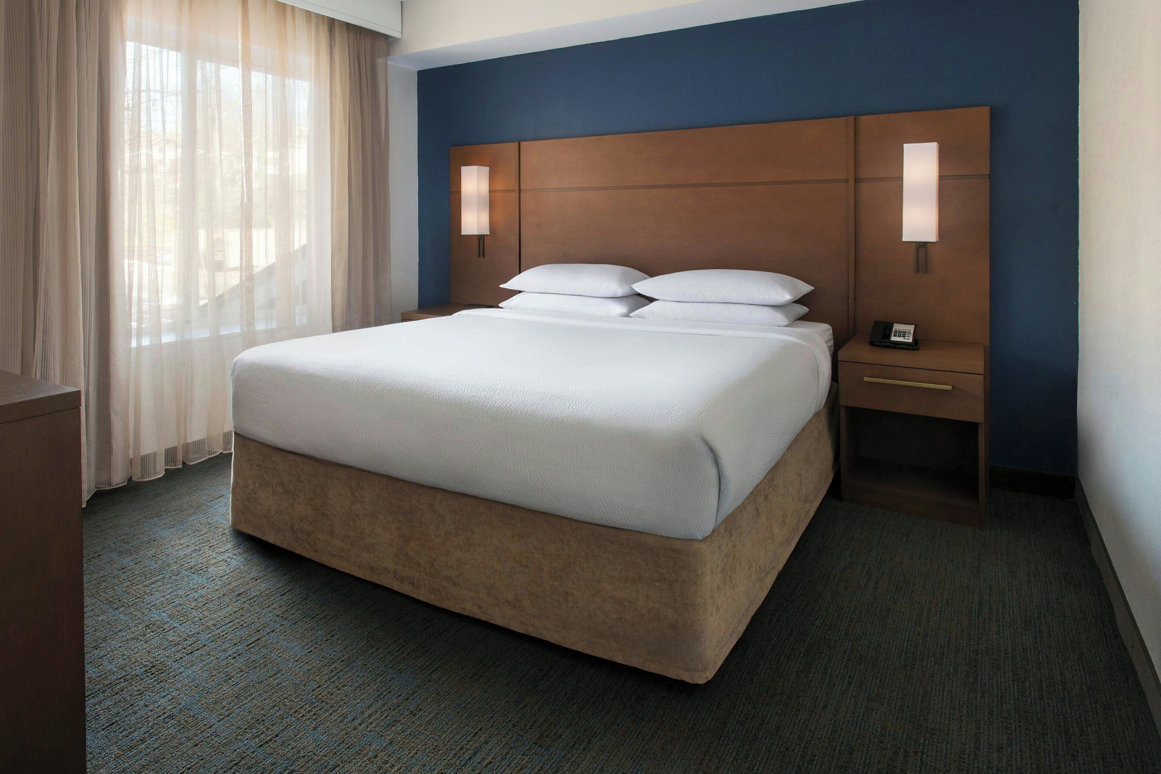 Quarto de hotel com uma cama grande, duas luminárias de cabeceira, mesa de apoio e uma janela com cortinas finas.