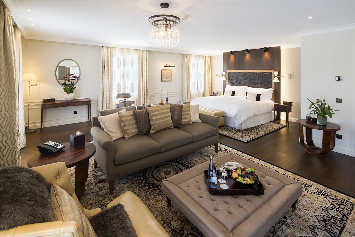 A modern hotel room with a large bed, a sitting area with a sofa and armchairs, a coffee table with fruit, and a chandelier.