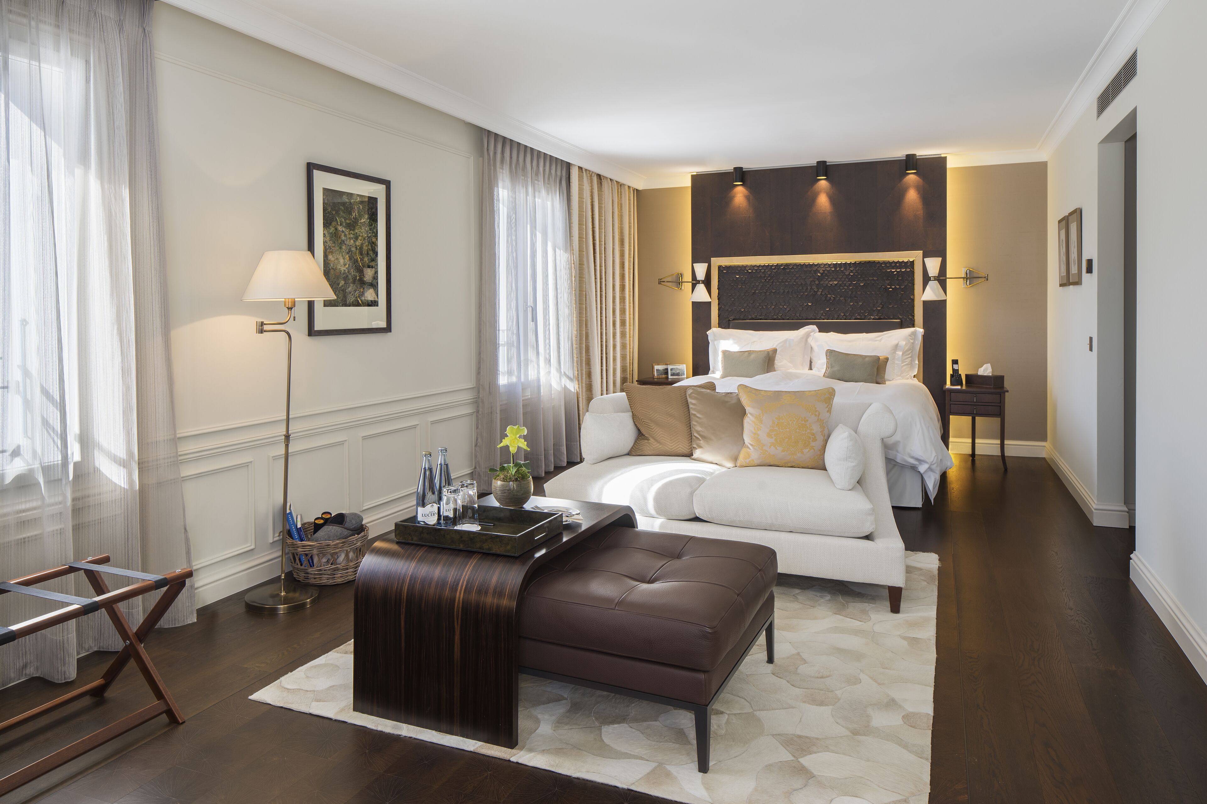 Una habitación de hotel moderna con una cama grande, una zona de estar con un reposapiés marrón, una mesita de noche con una lámpara y vistas a la ciudad.