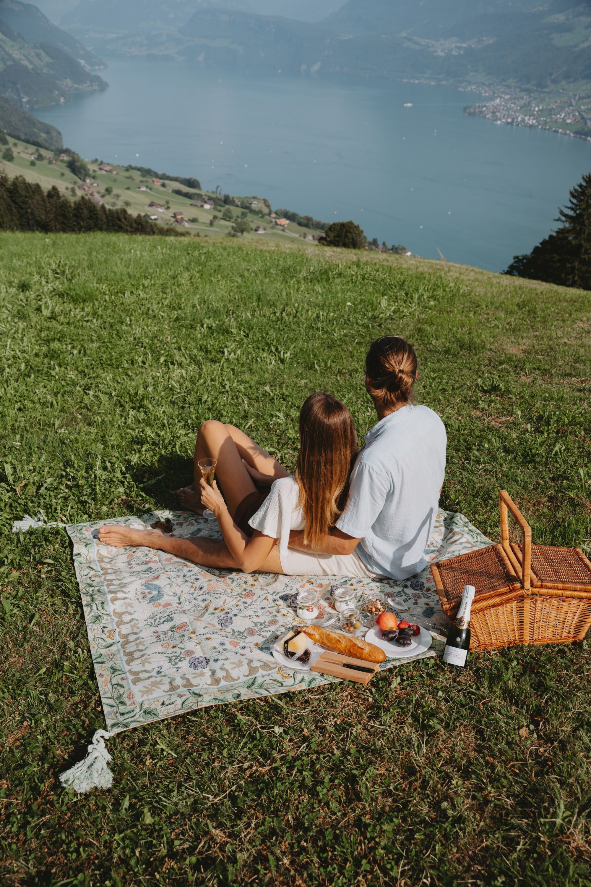 Pariskunta istuu piknik-vilttiin nojaten, ja heillä on piknik-tarvikkeet valmiina. He katselevat maisemia, johon kuuluu järvi ja kukkulat.