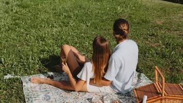 Pariskunta istuu piknik-vilttiin nojaten, ja heillä on piknik-tarvikkeet valmiina. He katselevat maisemia, johon kuuluu järvi ja kukkulat.