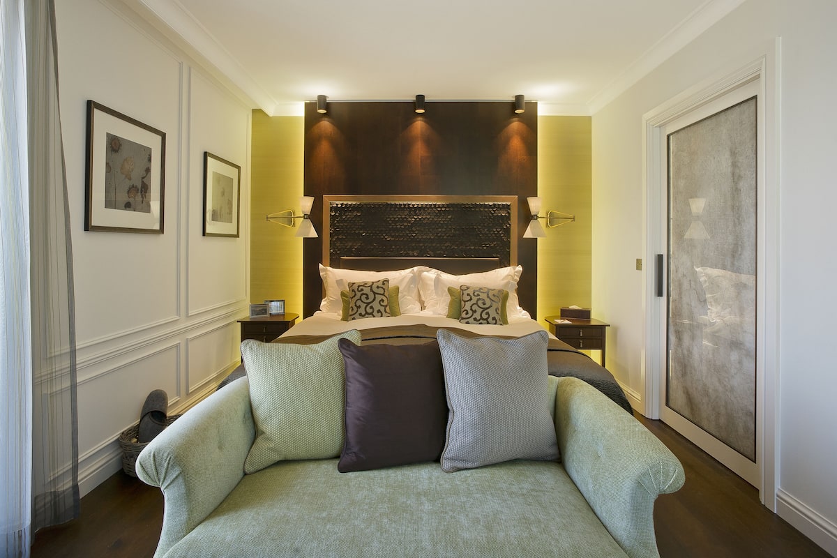 A bedroom with a large bed, a headboard with built-in lighting, a sofa with multiple cushions, and framed artwork on the walls.