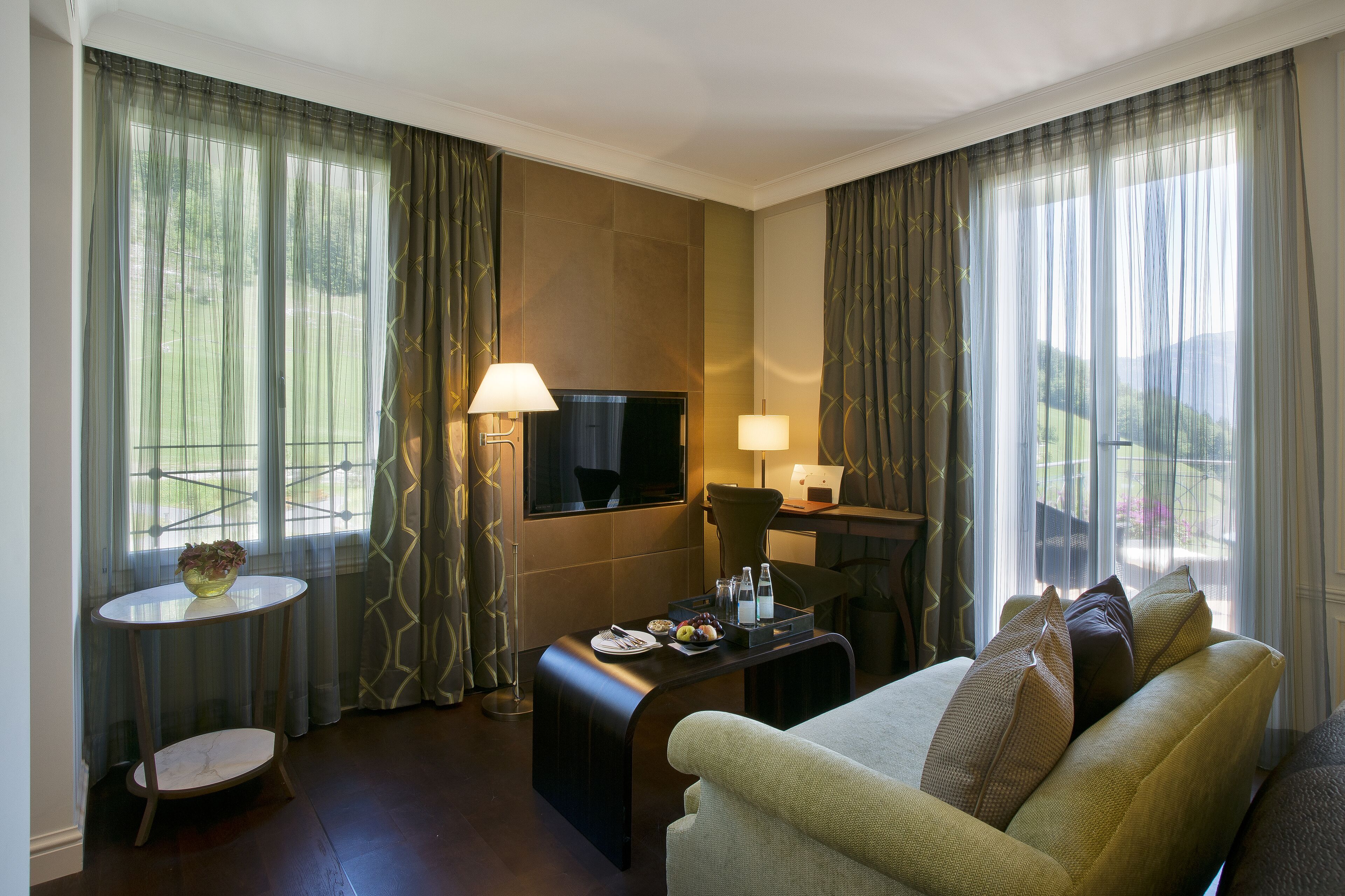 A hotel room with a sofa, a small table with a bowl of fruit, a flat-screen TV, and a desk with a lamp.