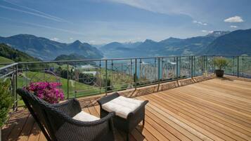 Eine Holzterrasse mit Korbstühlen und einem Blumentopf, mit Blick auf eine Berglandschaft und einen See.