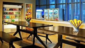 A modern dining area with long tables and benches, a refrigerated beverage display, and a large window with blue curtains. - Aloft Zhengzhou Shangjie (Zhengzhou)