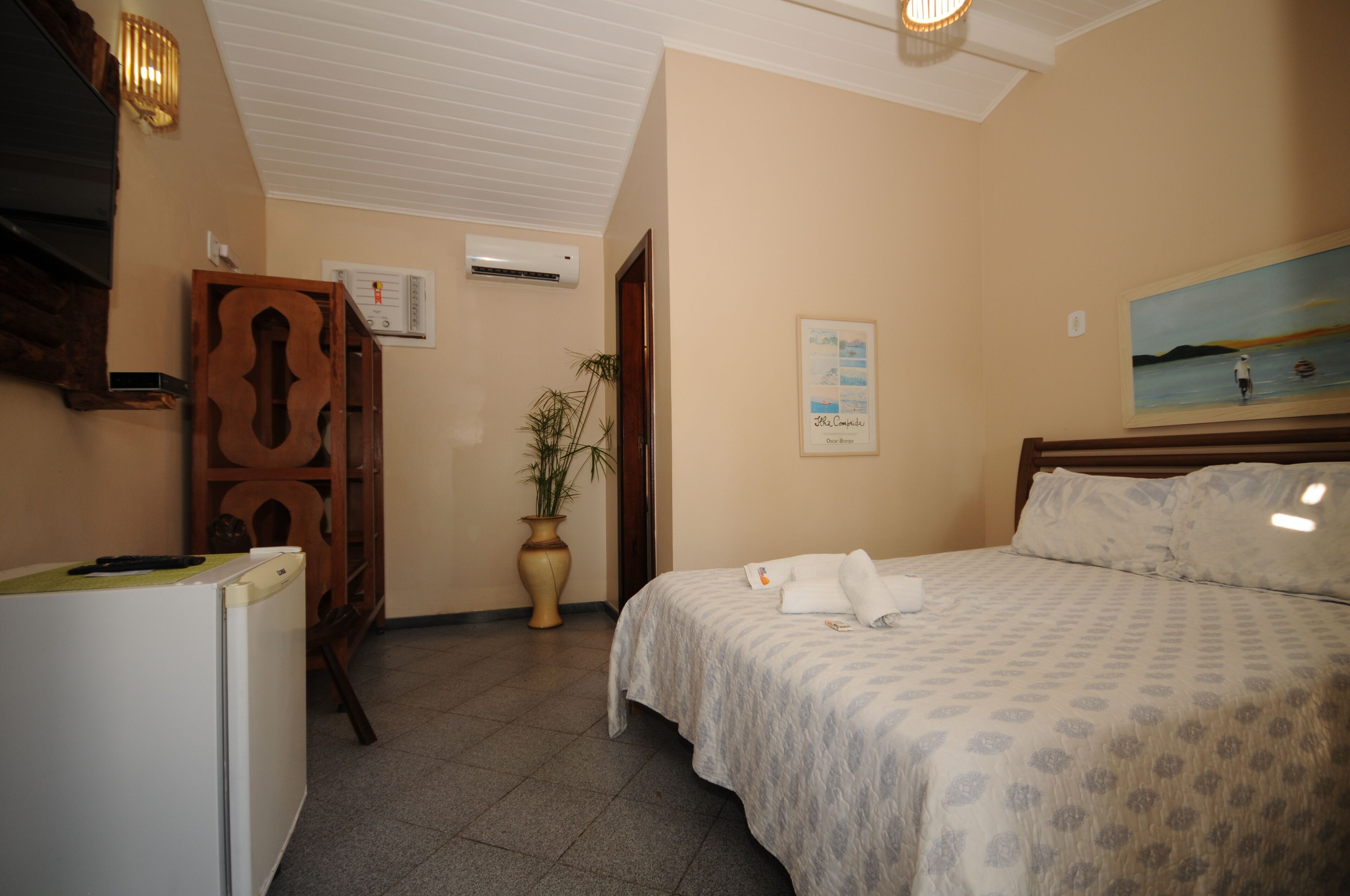 Quarto de hotel com cama, escrivaninha, frigorífico, planta em vaso e um quadro na parede.