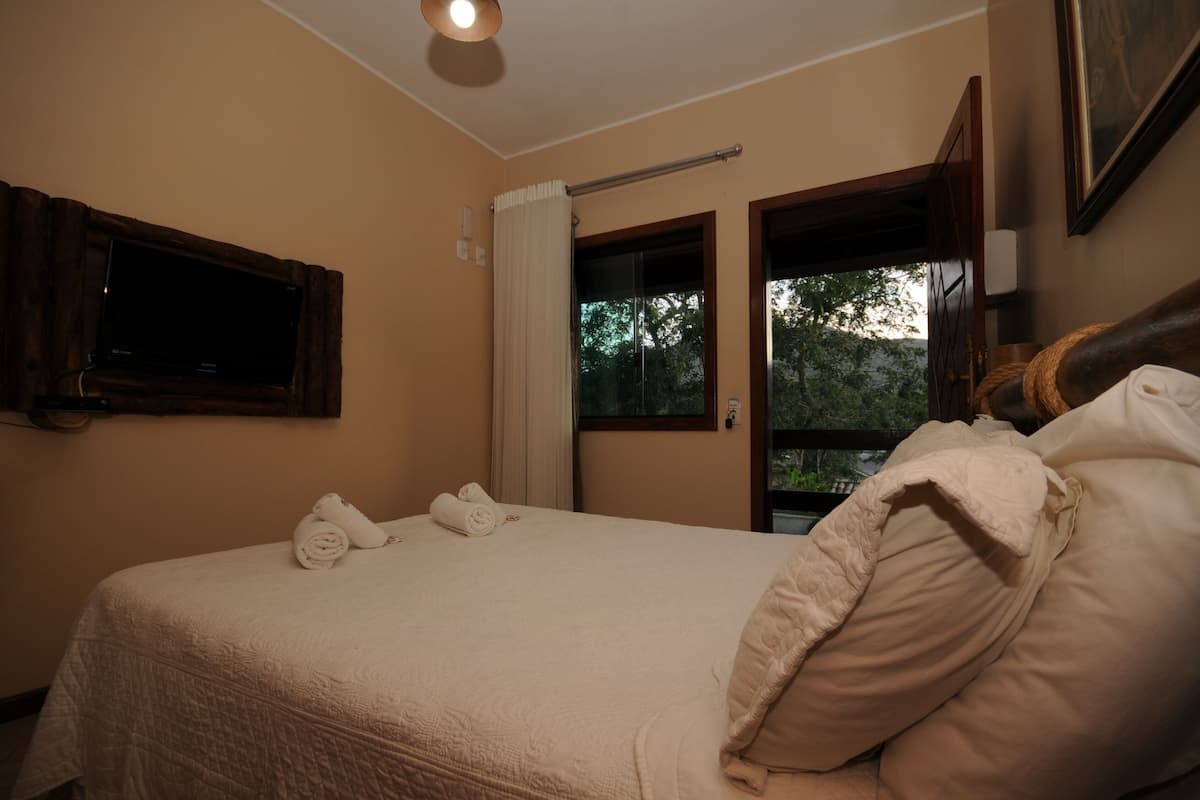 A hotel room with a bed, a television mounted on the wall, a window with curtains, and a view of trees outside.
