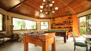 Ein großzügiger Innenbereich mit Kicker-, Billard- und Pooltisch. Korbmöbel und ein Esstisch mit Stühlen sind ebenfalls vorhanden.