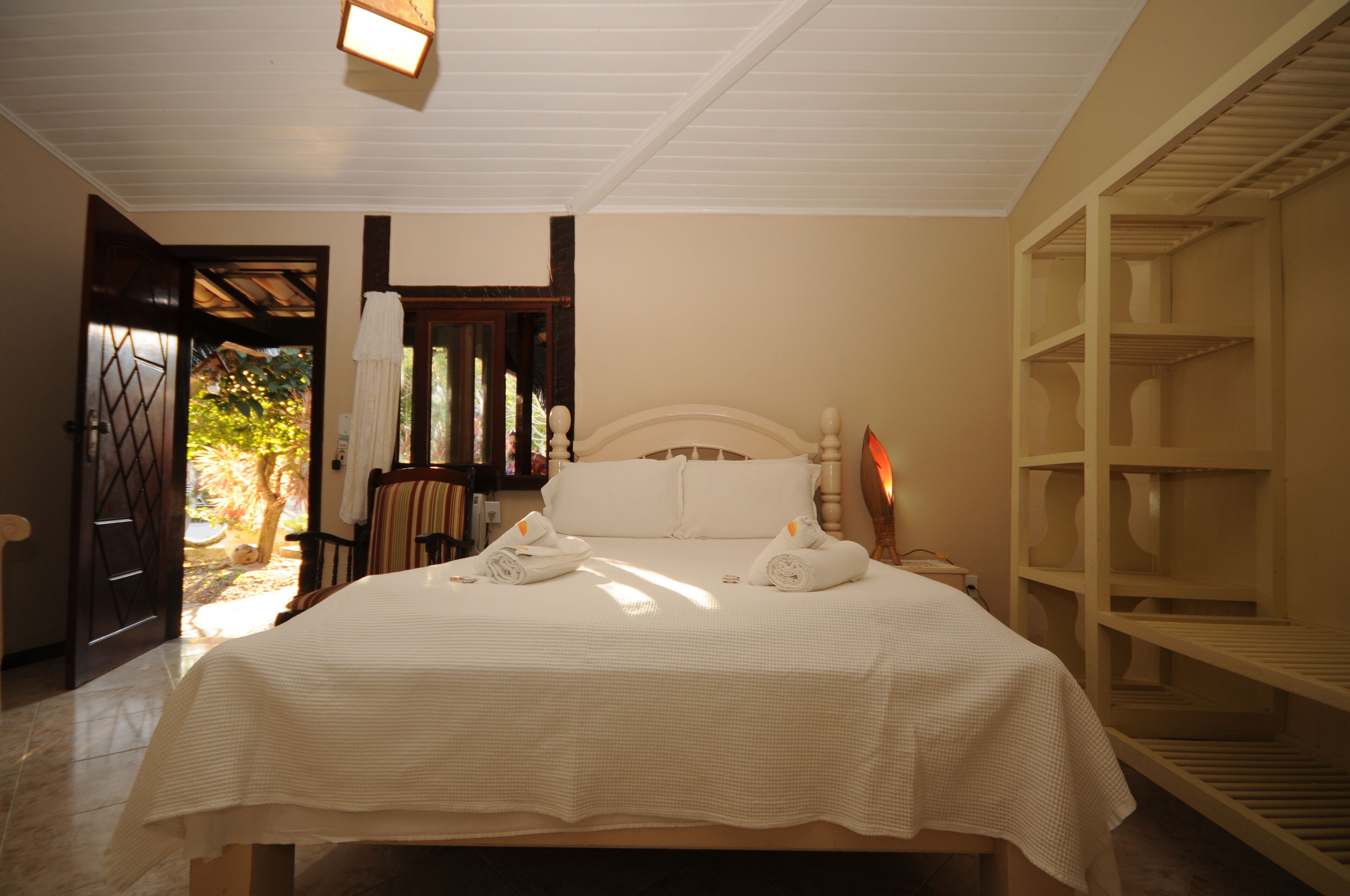 Cama bem arrumada com lençóis e toalhas brancas, cabeceira de madeira e uma estante à direita.