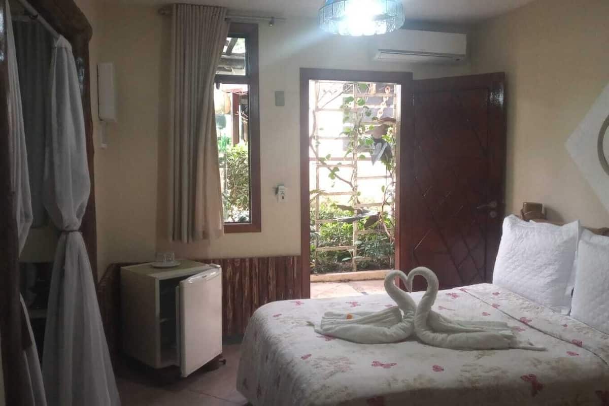 A bedroom with a bed, a small table, a refrigerator, a window, and a door leading to an outdoor area.