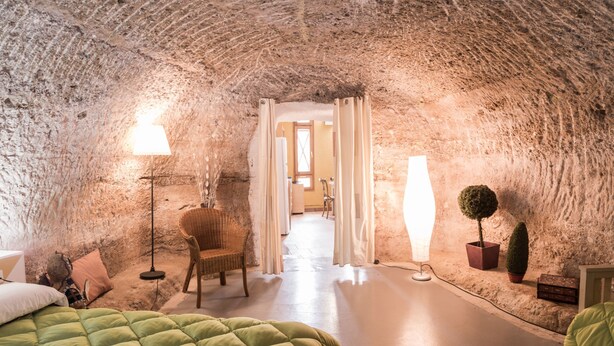 Una habitación con techo de piedra, una cama con colcha verde, una silla de mimbre, una lámpara de pie y una planta en maceta.