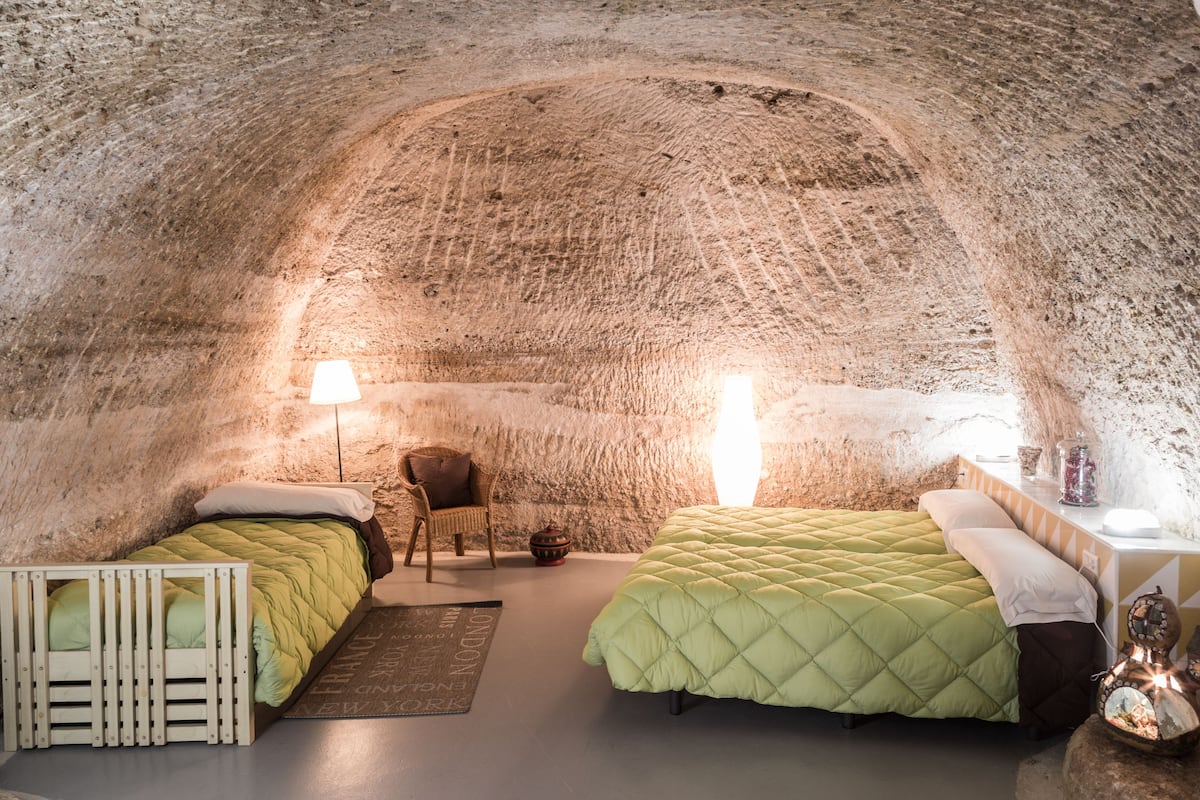 A cozy, rustic room with a vaulted ceiling, two beds with green quilts, a wicker chair, a small table with a lamp, and a decorative item on the table.