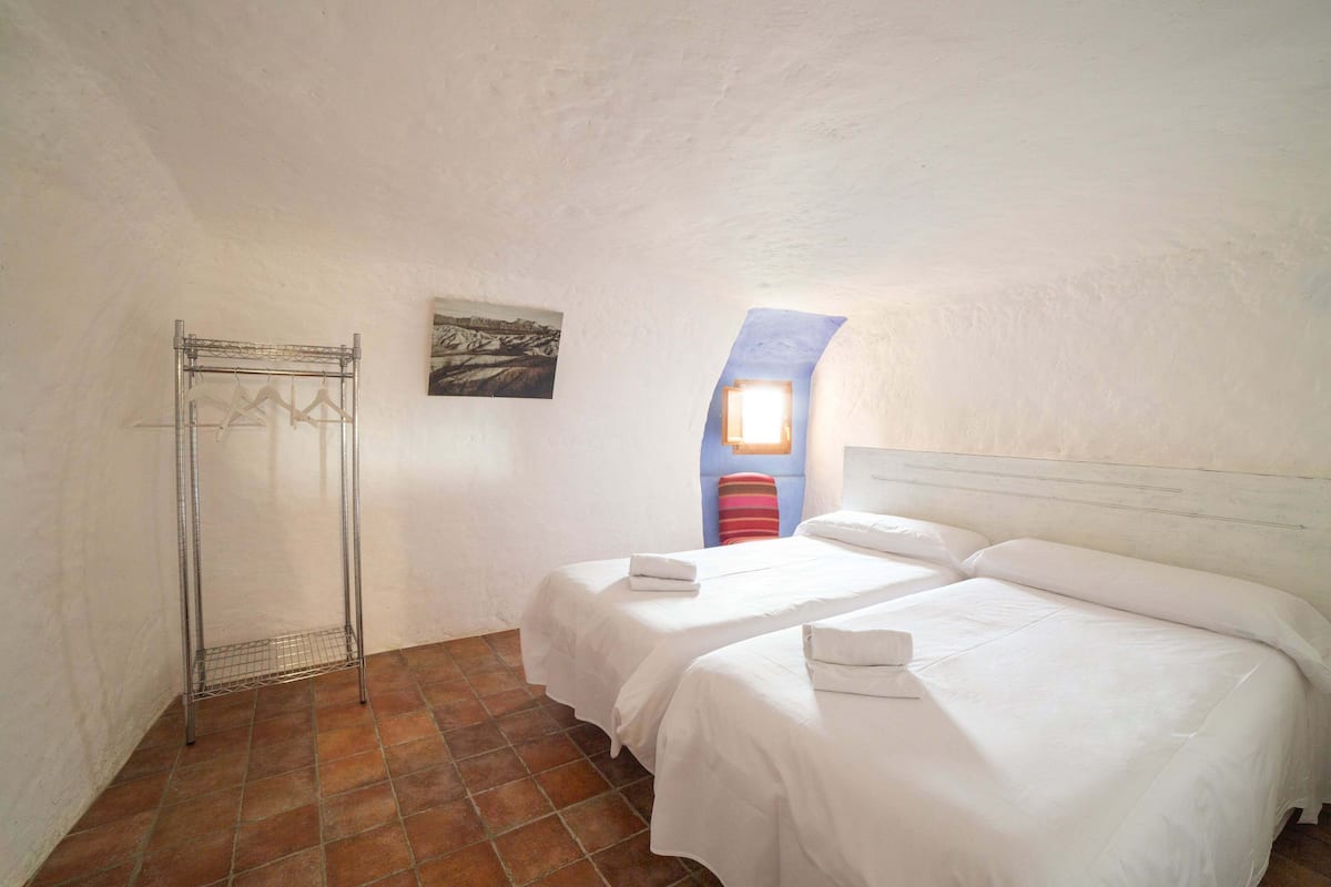 A room with two beds, a metal drying rack, a painting, and a window with a view.