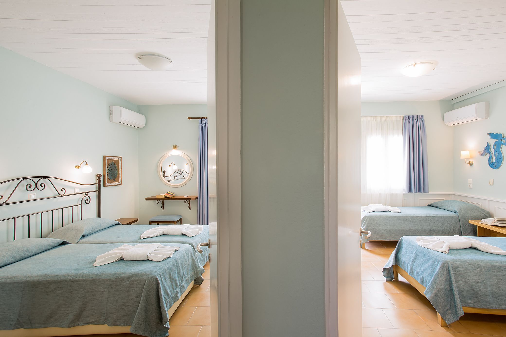 A hotel room with two single beds, a small table, a mirror, and a window with curtains.