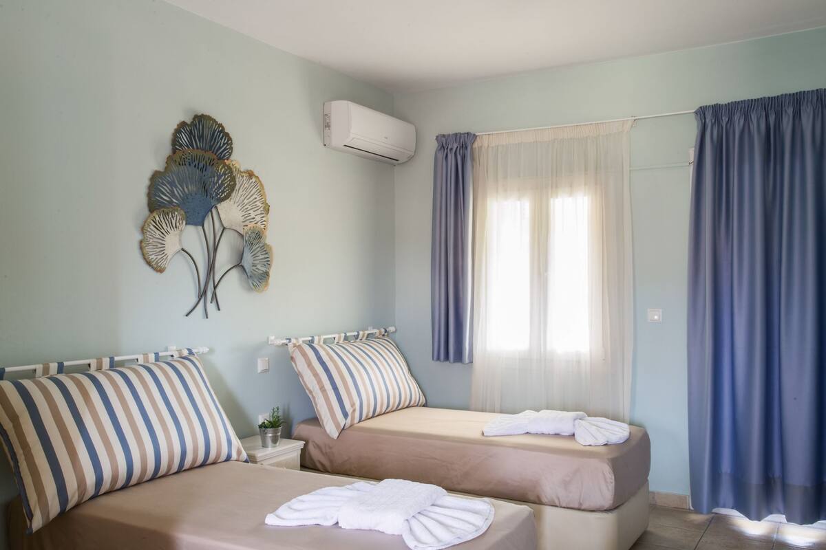 A hotel room with two beds, a wall-mounted air conditioner, and a window with blue curtains.