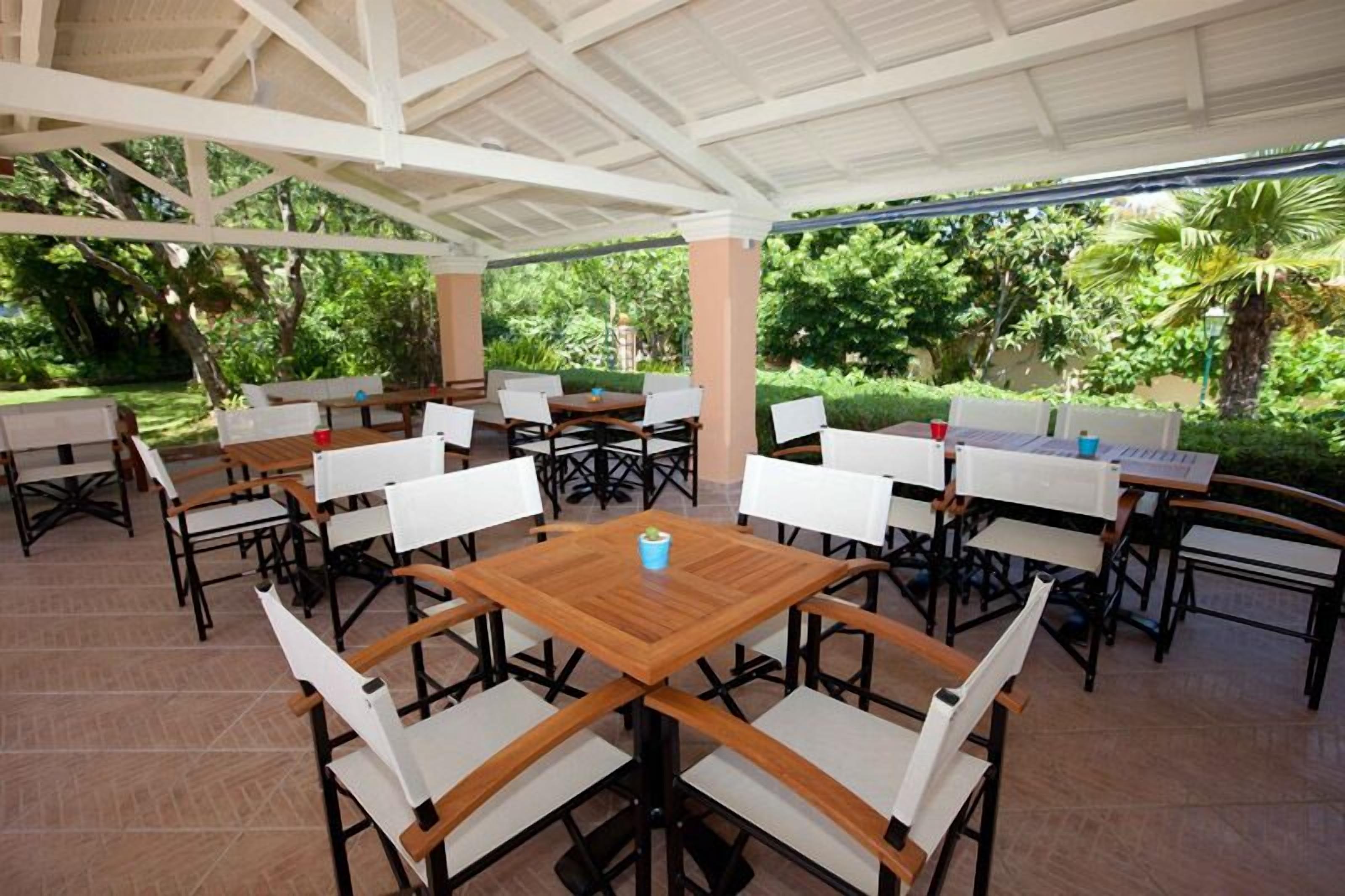 An outdoor covered dining area with multiple tables and chairs, surrounded by lush greenery.