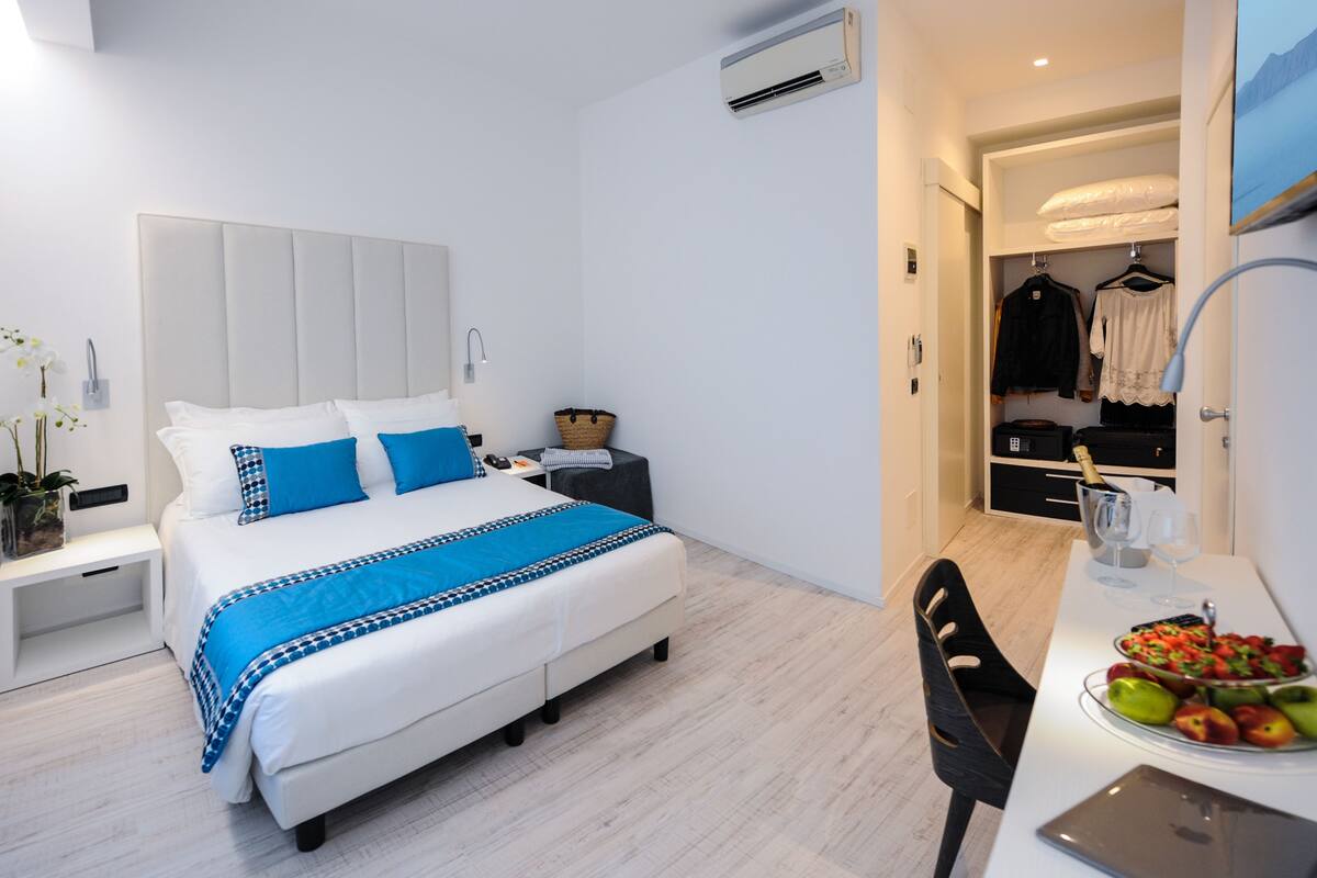 Une chambre d’hôtel moderne avec un grand lit, des tables de chevet, une petite table avec des fruits et une armoire où sont suspendus des vêtements.