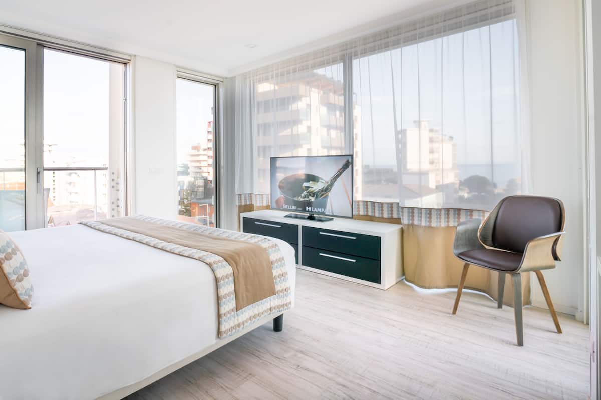 Une chambre d’hôtel avec un grand lit, un bureau avec une télévision et une vue sur la ville grâce à de grandes fenêtres.