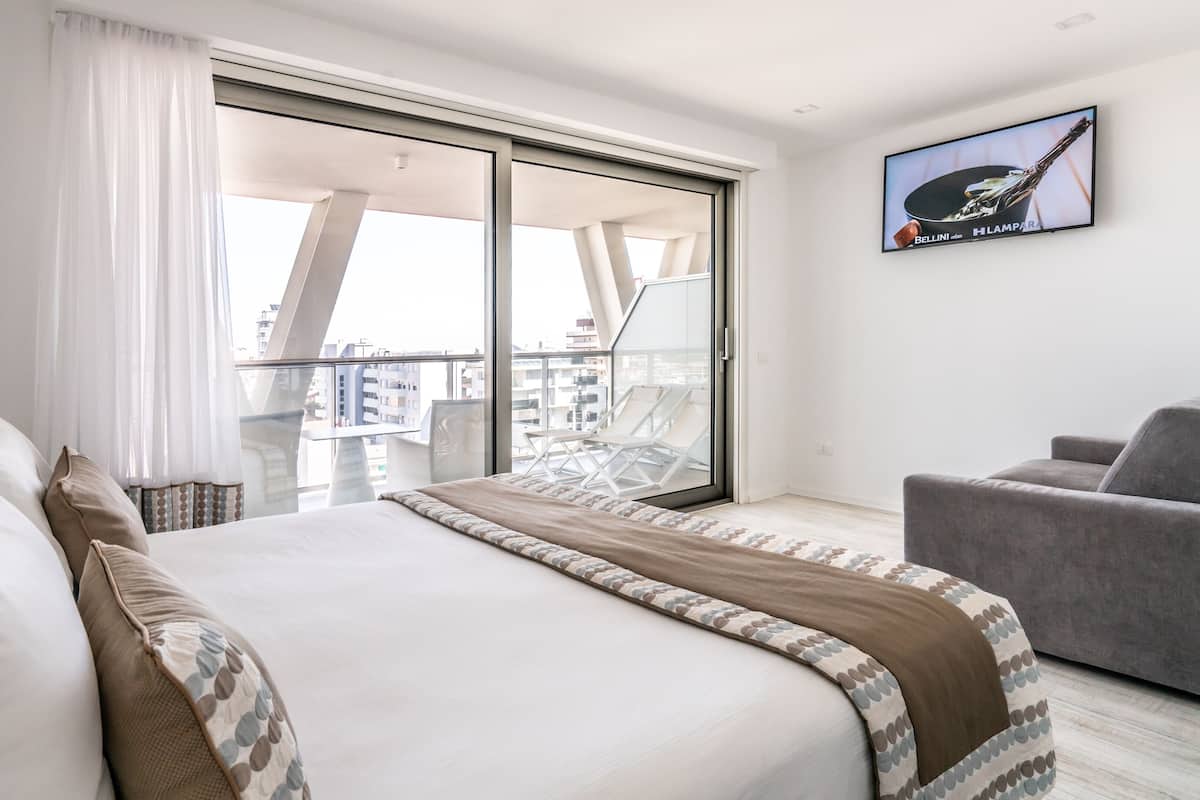 Une chambre d’hôtel moderne avec un grand lit, un canapé gris, un balcon offrant une vue sur la ville et un téléviseur fixé au mur.