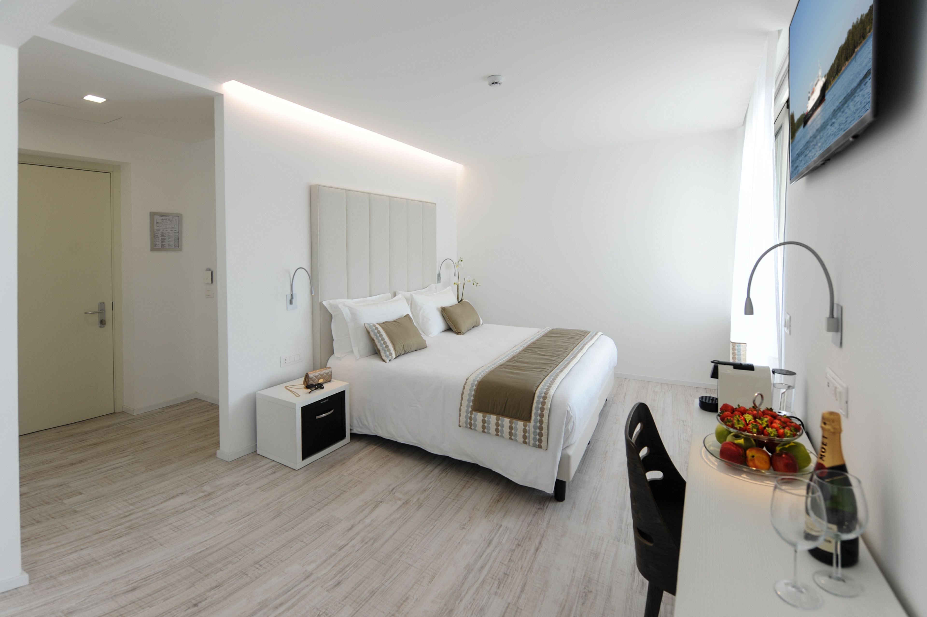 A modern bedroom with a bed, bedside table, and a small dining area with a fruit bowl and wine glasses.