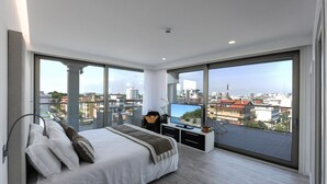 Een moderne slaapkamer met een groot bed, een televisie en uitzicht op de stad door middel van kamerhoge ramen.