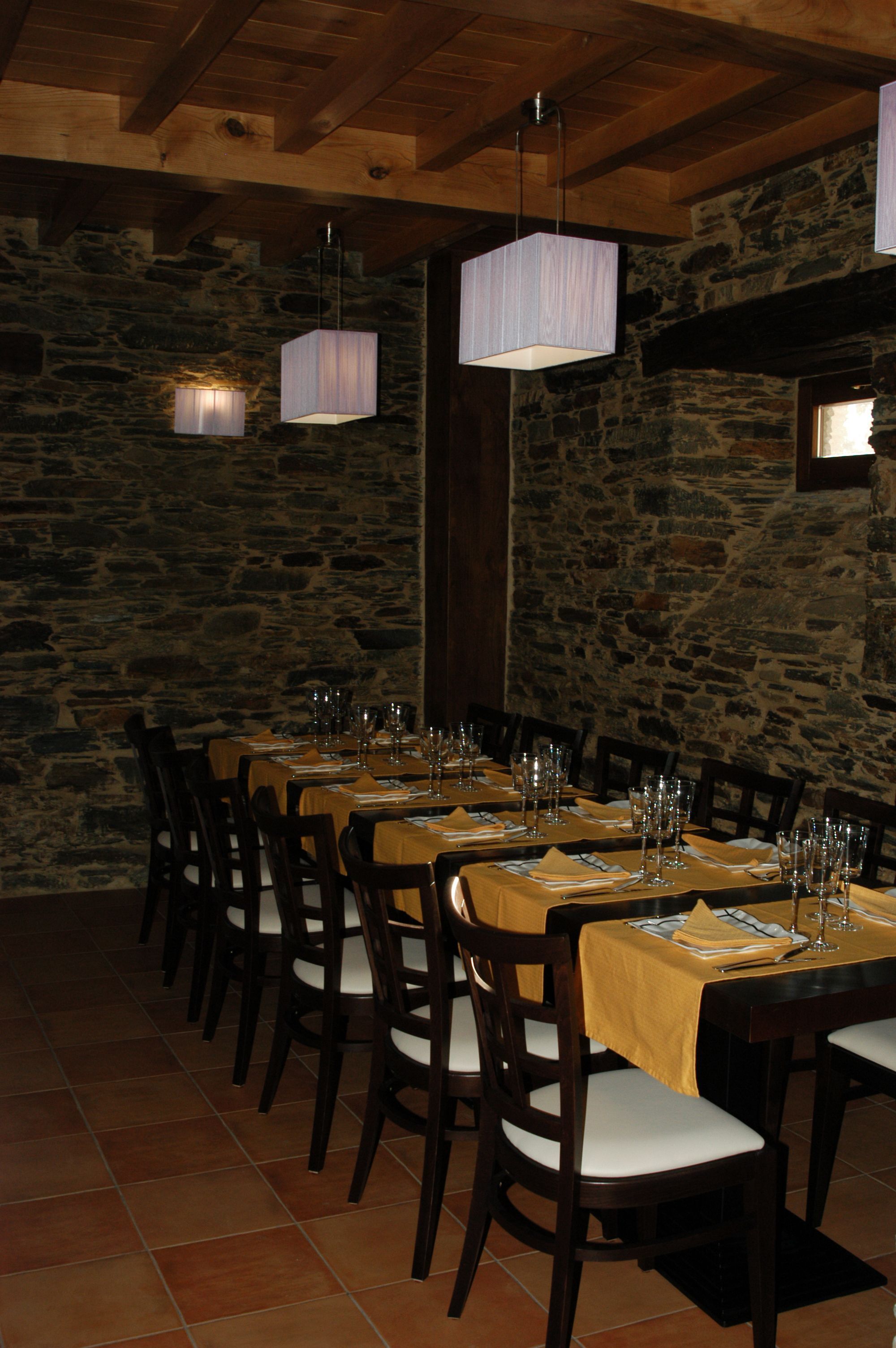 A long dining table set with chairs, wooden beams, stone walls, and hanging light fixtures.