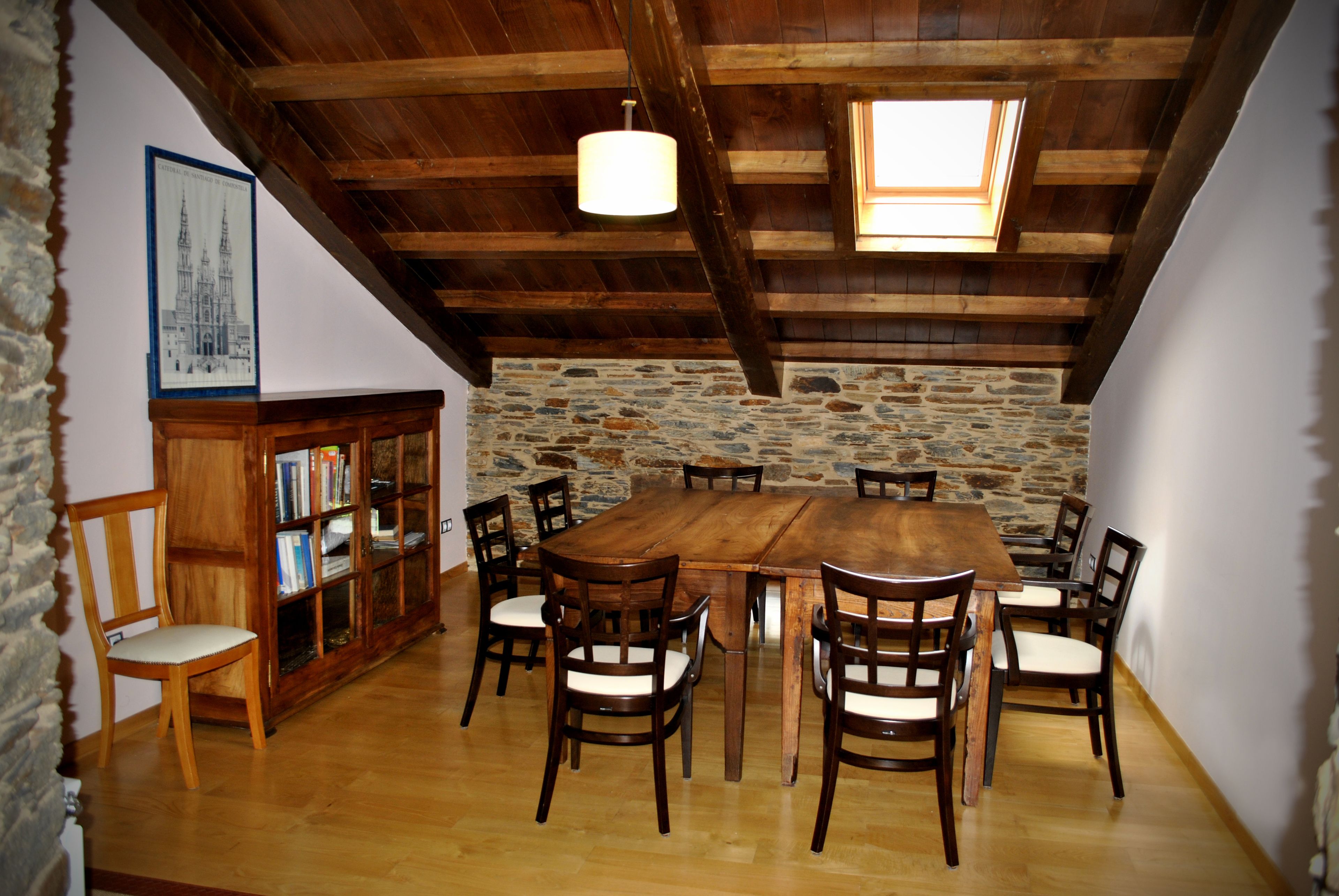 A dining area with a large wooden table surrounded by chairs, a stone wall, and a wooden bookshelf with books.