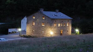 A stone house with multiple windows and a chimney, surrounded by a grassy field and trees.