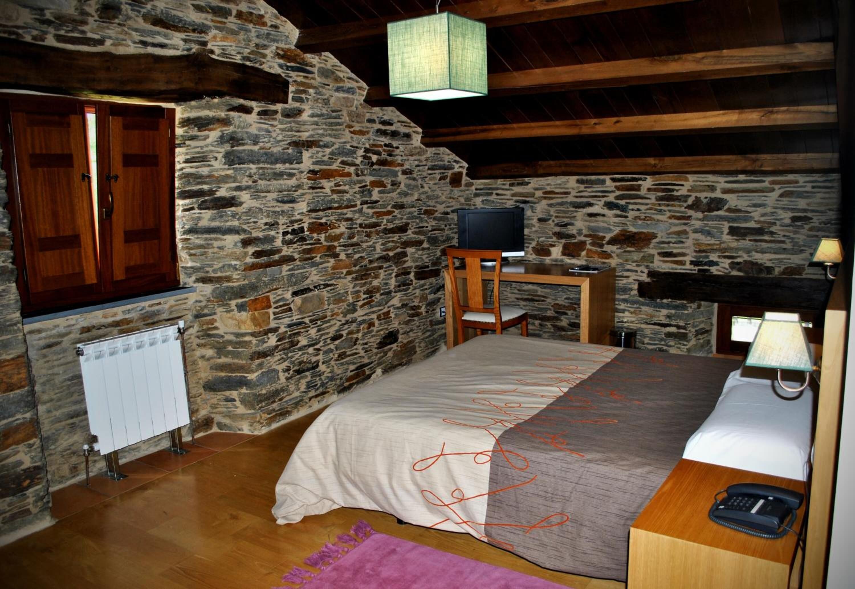 Una camera rustica con parete in pietra, travi in legno, un letto con copriletto beige e grigio, un comodino in legno con una lampada, una sedia e una televisione su un tavolino.