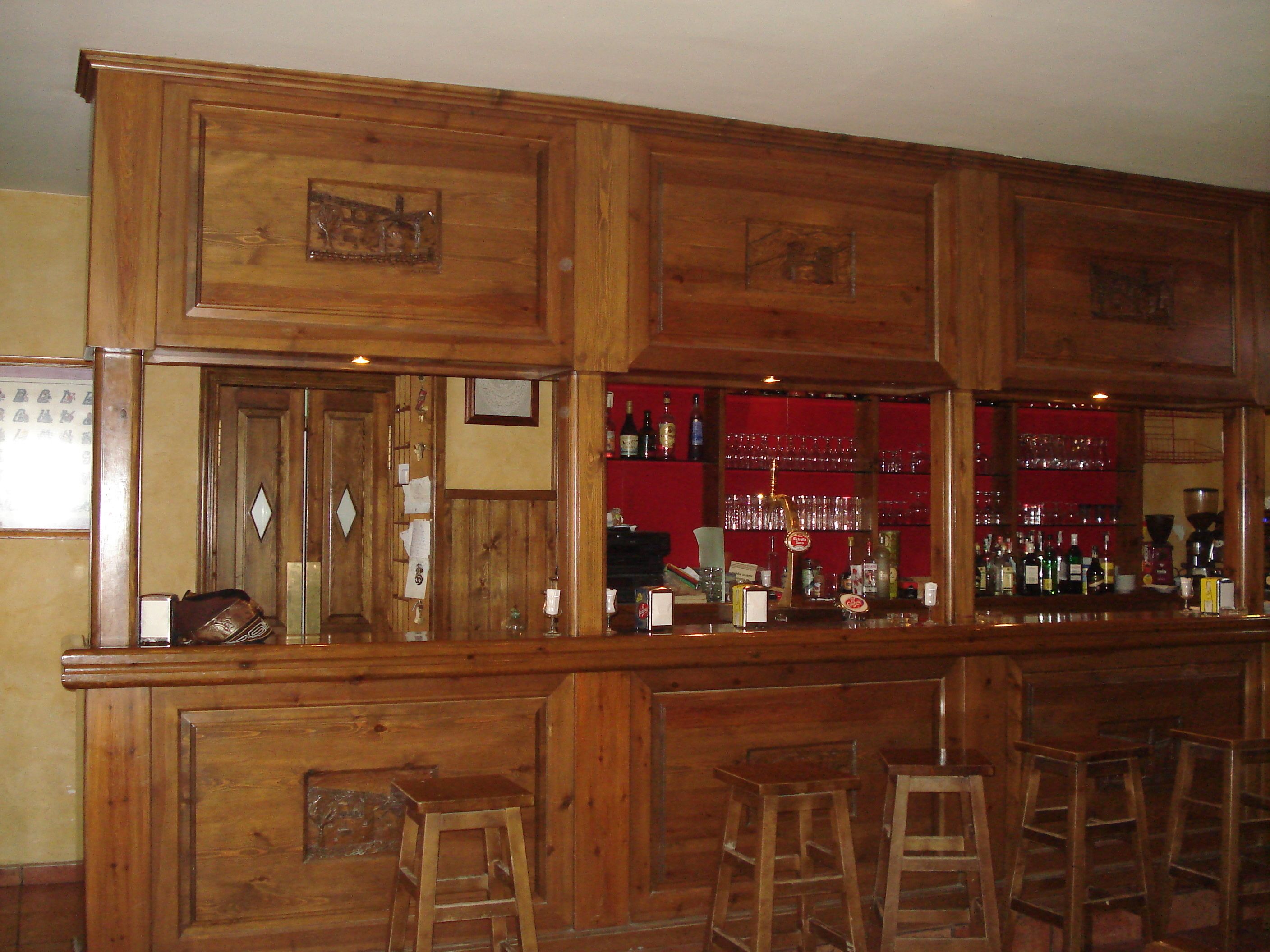 Una barra de madera con taburetes, un fondo rojo y diversas botellas y vasos.