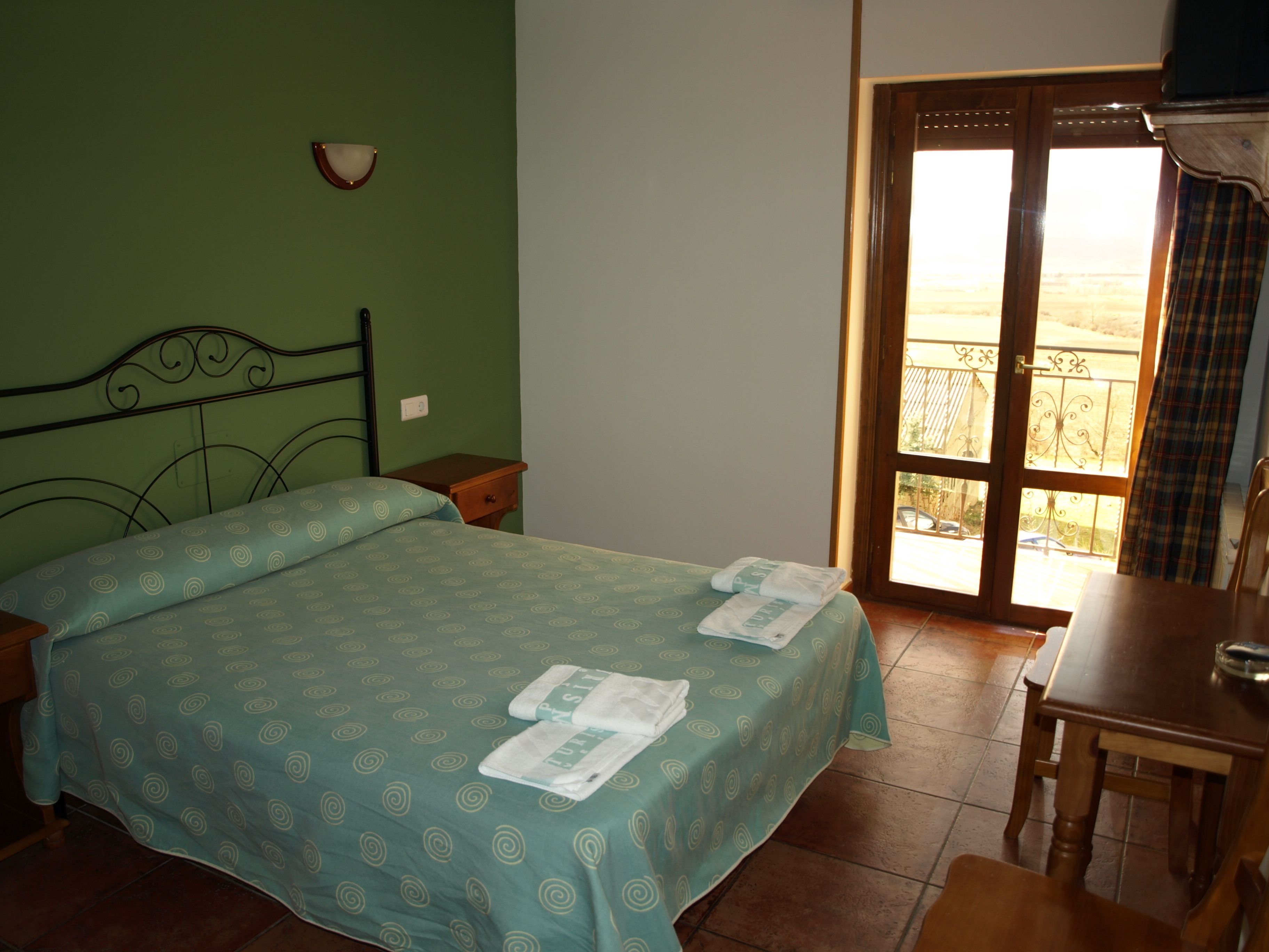 A bedroom with a bed, a nightstand, a wooden table, and a balcony with a view.