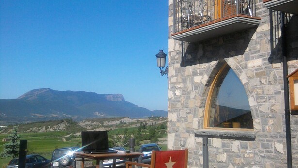 A stone building with a balcony, a table with chairs, and a view of mountains. - Hotel El Churron (Sabinanigo)