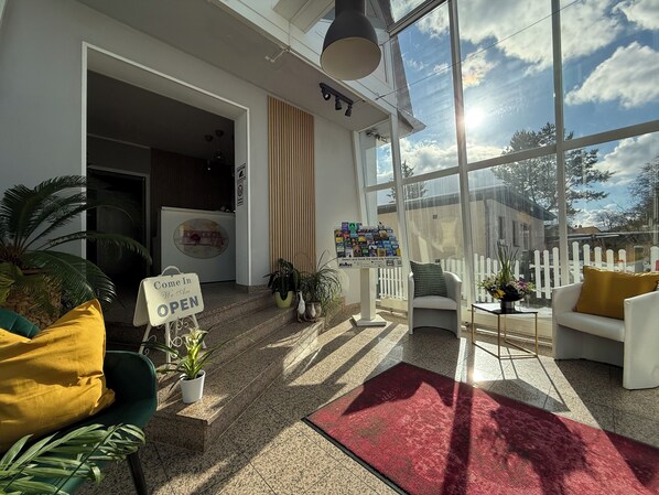 A hotel lobby with a red carpet, a 'Come In We Are OPEN' sign, a reception desk, and a large window offering a view of the sky and trees.
