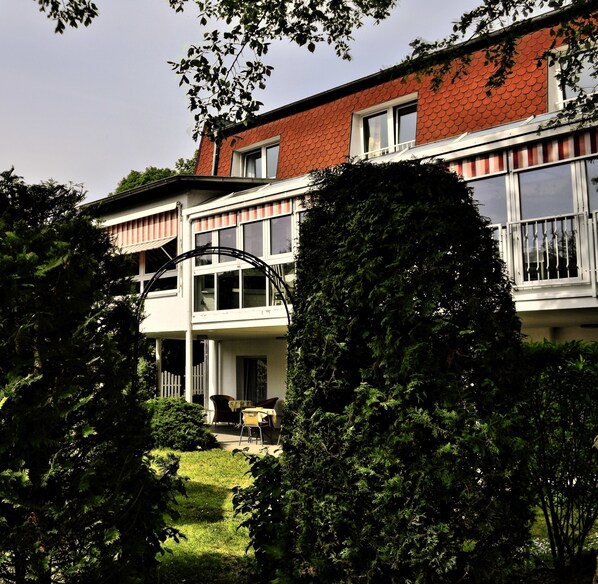 A two-story building with a red roof, white walls, and large windows. There is a covered outdoor seating area with chairs and a table.