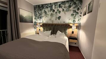 Une chambre d’hôtel avec un lit, une tête de lit ornée d’un motif de feuilles, deux tables de chevet avec des lampes et un tableau encadré au mur.
