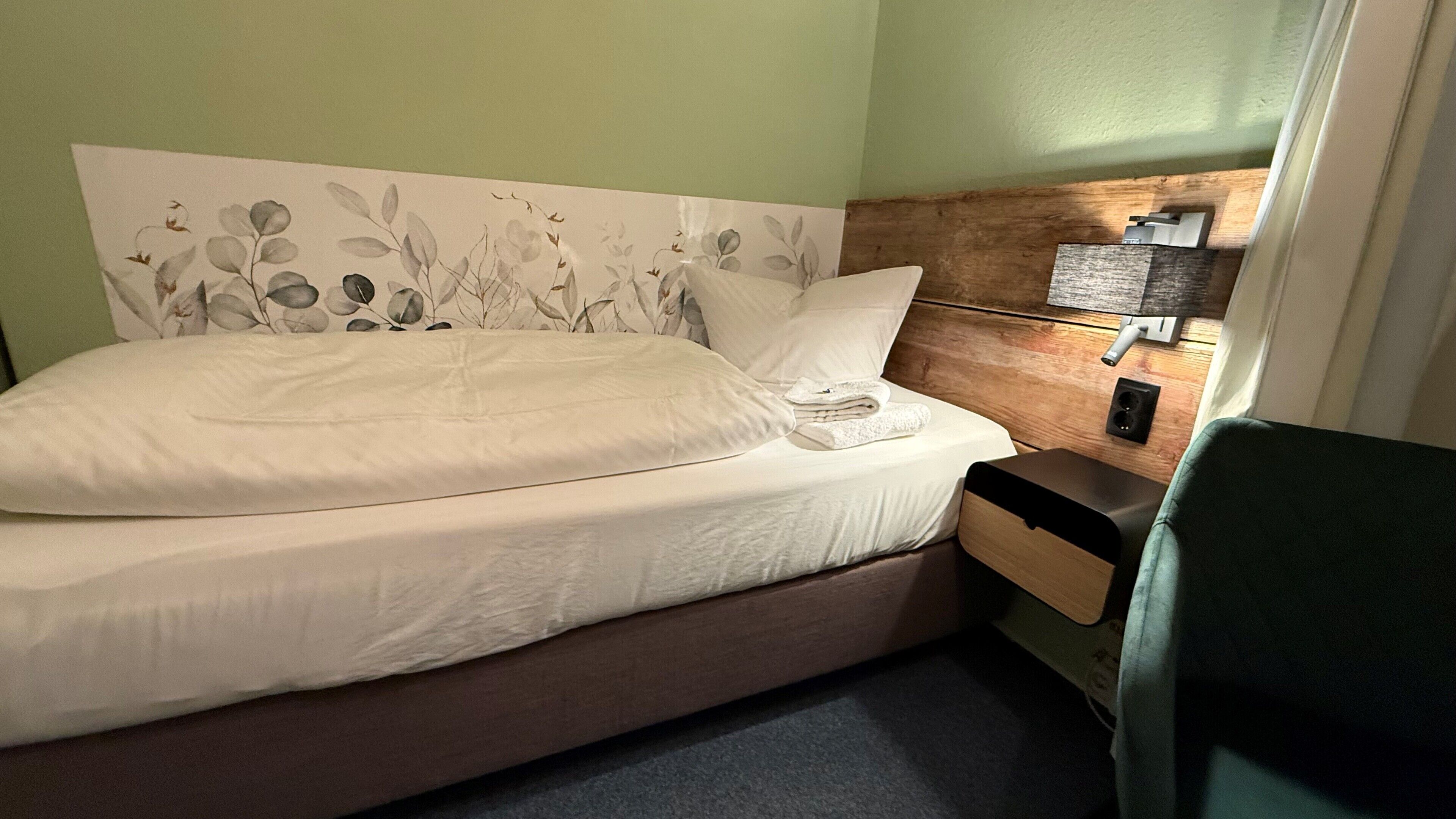 Een net opgemaakt bed met een bloemrijk hoofdeinde, een nachtkastje met een lamp en een groene stoel.