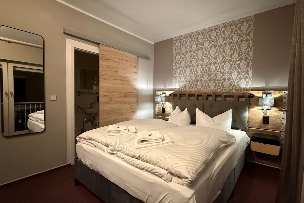 A hotel room with a large bed, a wooden headboard, a mirror, a bathroom visible through an open door, and a patterned wallpaper.