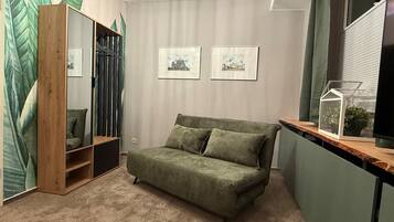 A room with a green sofa, a wooden wardrobe, two framed pictures on the wall, and a small plant in a glass container on a wooden shelf.