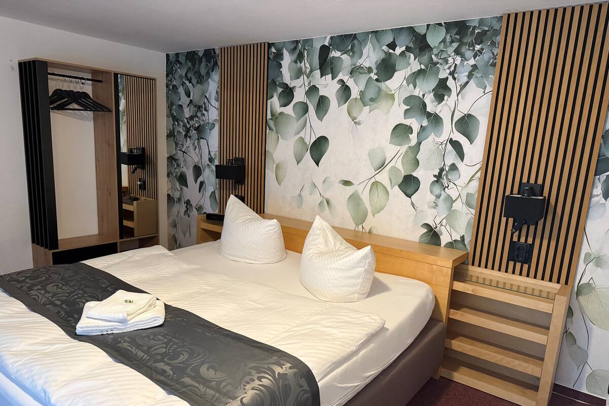 A neatly made bed with white linens and a black patterned throw, a wooden headboard, and a wall with a leaf pattern.