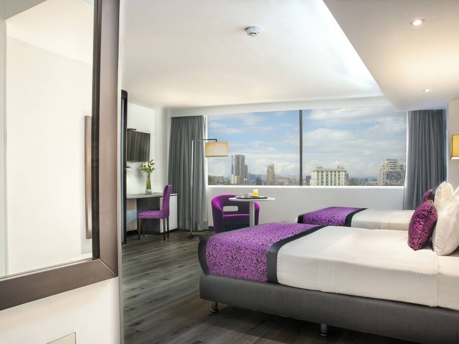 Habitación de hotel con una cama grande, un escritorio, una silla y vistas a la silueta del horizonte urbano.