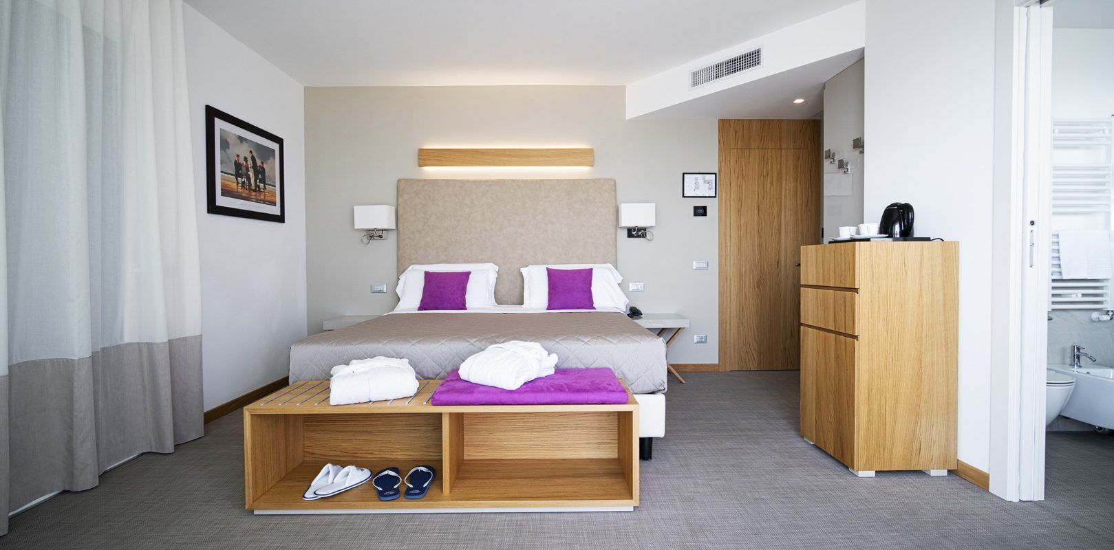 A modern hotel room with a large bed, a wooden desk, a wardrobe, and a bathroom visible through an open door.
